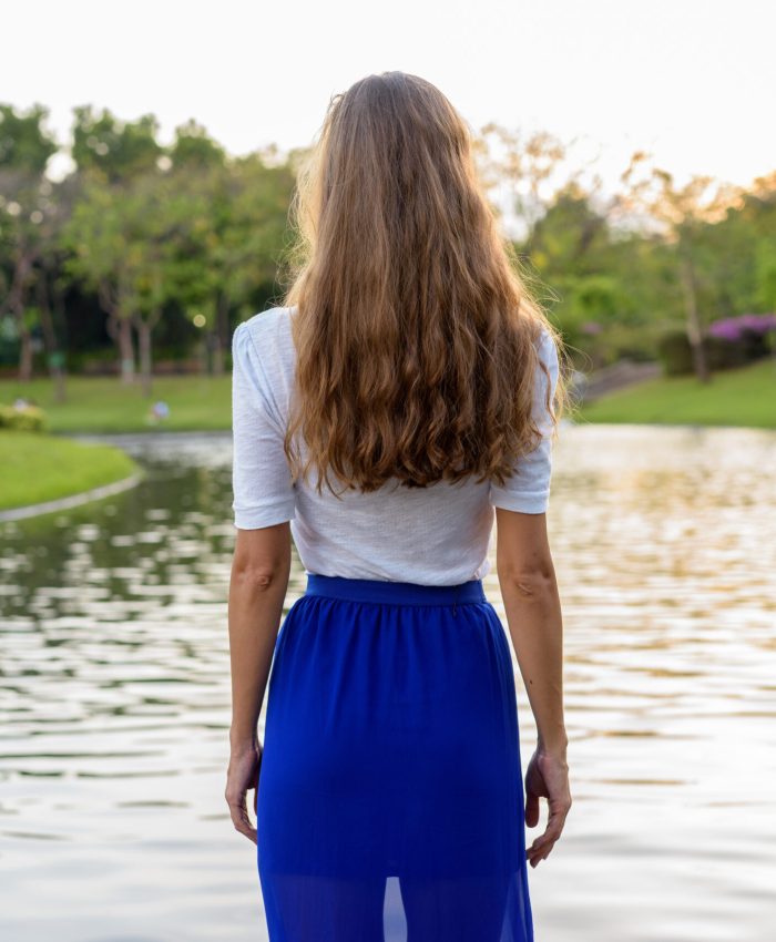 Beautiful woman with long hair standing and looking at scenic view of the lake in peaceful green park